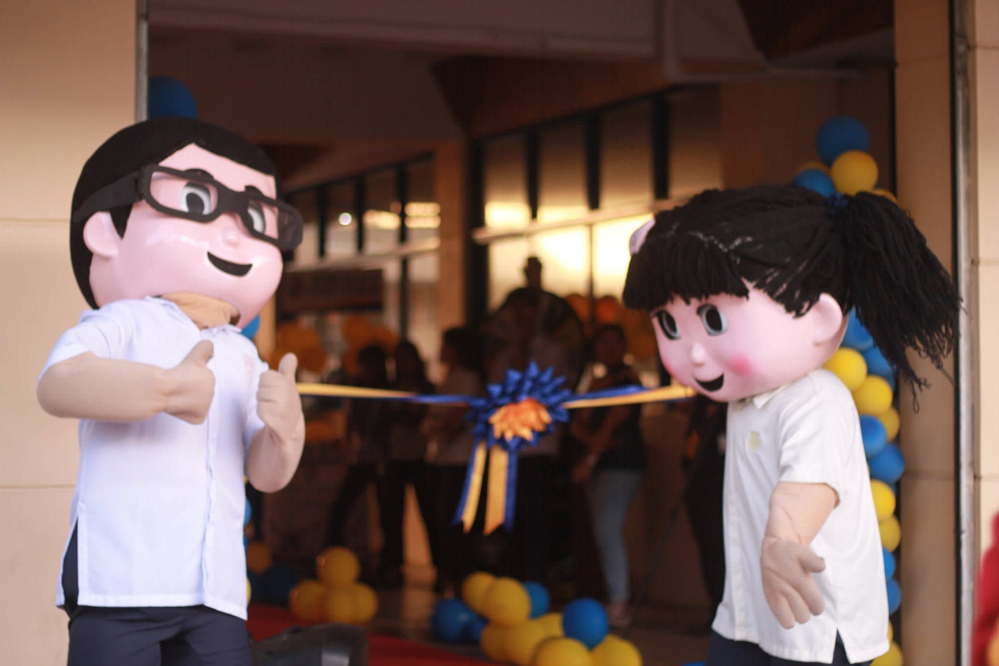 Two student mascots in white shirts greet visitors at a campus opening marked by blue and gold balloons